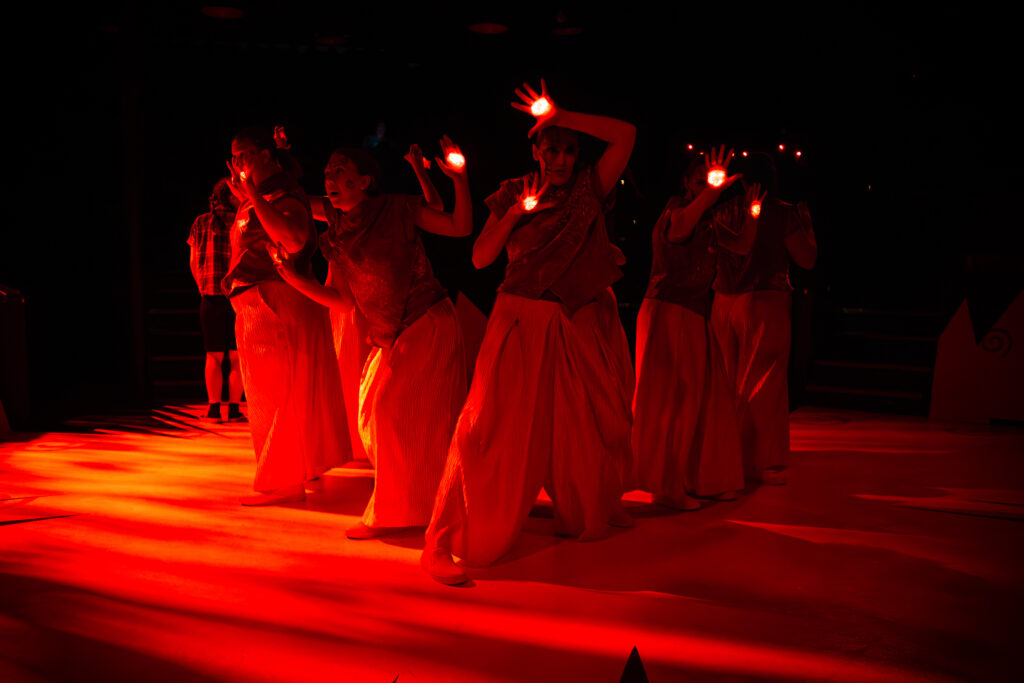The acting ensemble, in matching white loose pants, and gray space-aged shirts have red glowing eyes on their hands. The entire stage is bathed in red light.